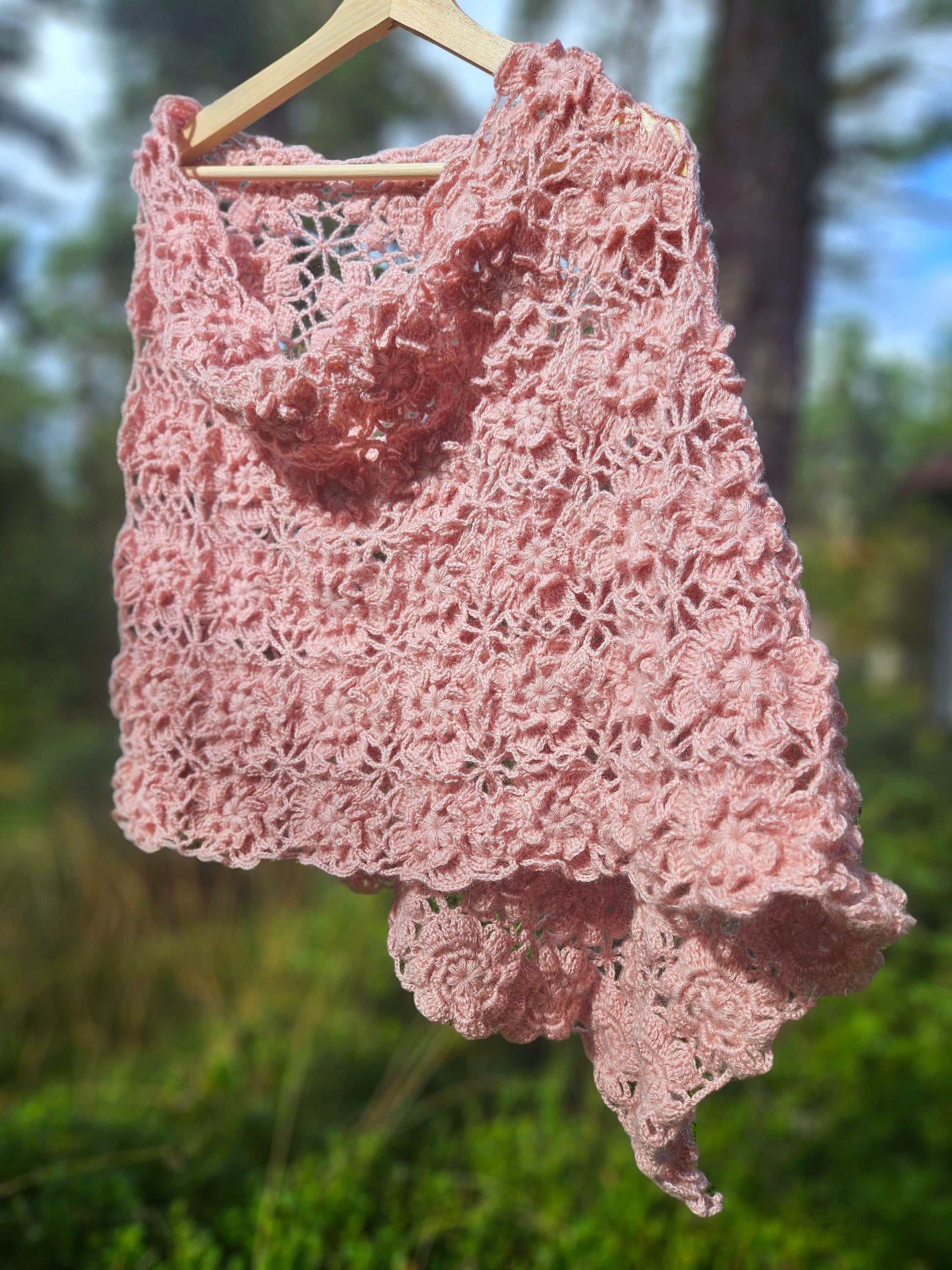 Pink crochet sakura poncho hanging on a wooden hanger against a blurred natural background
