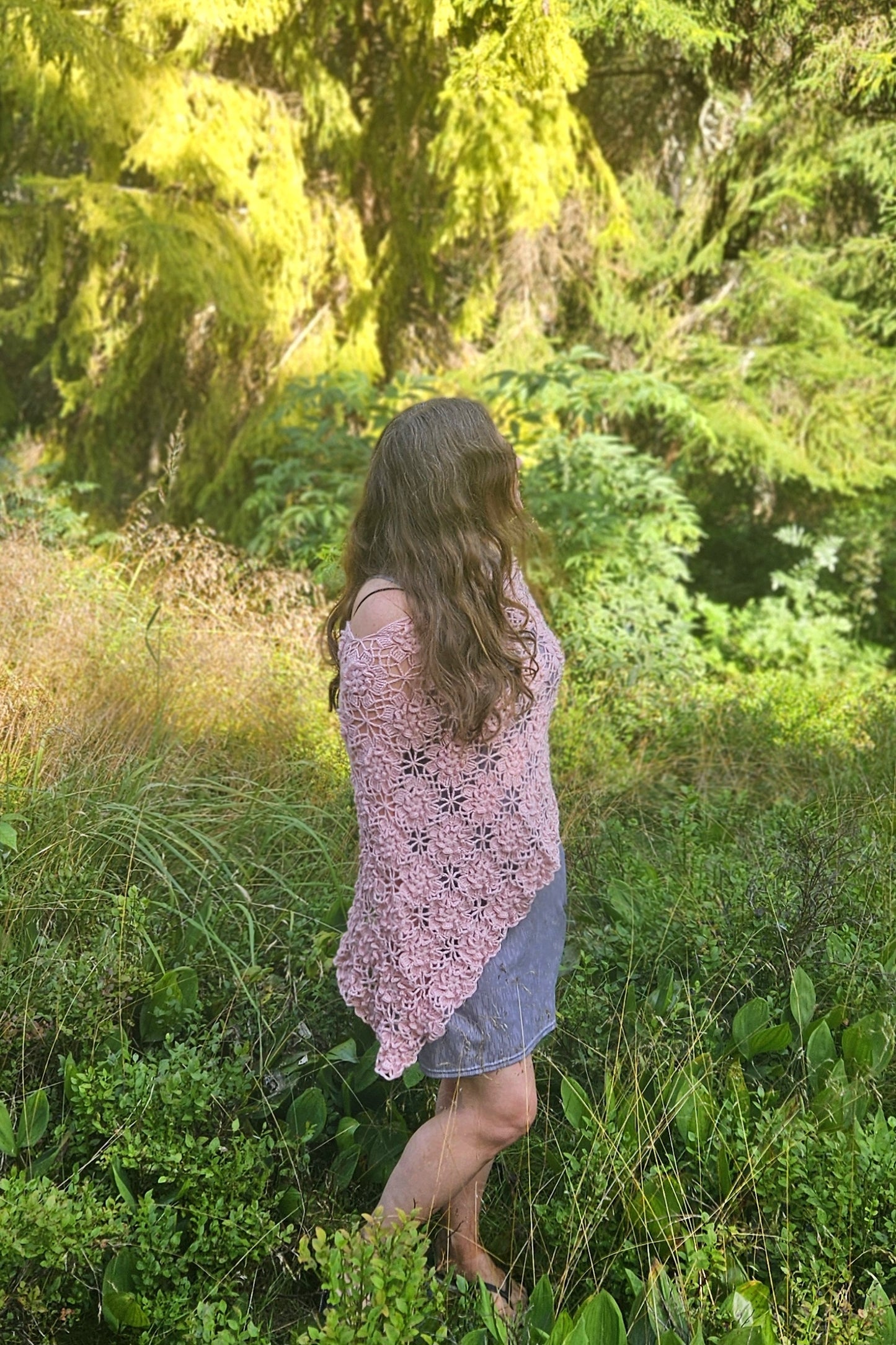 Woman in a pink crochet sakura poncho standing in a lush green forest