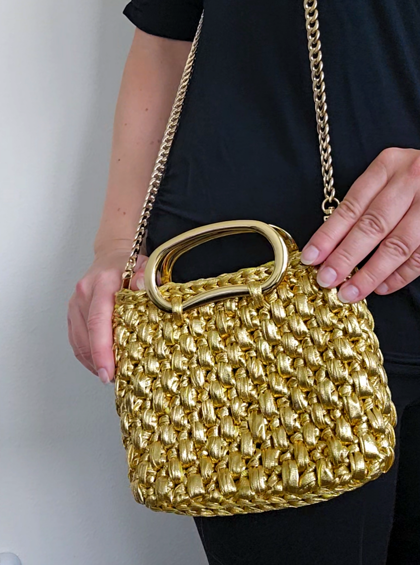 Gold crochet handbag with a chain strap held by a person against a neutral background
